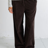 A person wearing dark brown corduroy wide-leg trousers, a cream and navy striped long-sleeve top, and black pointed flats, standing against a white textured wall.