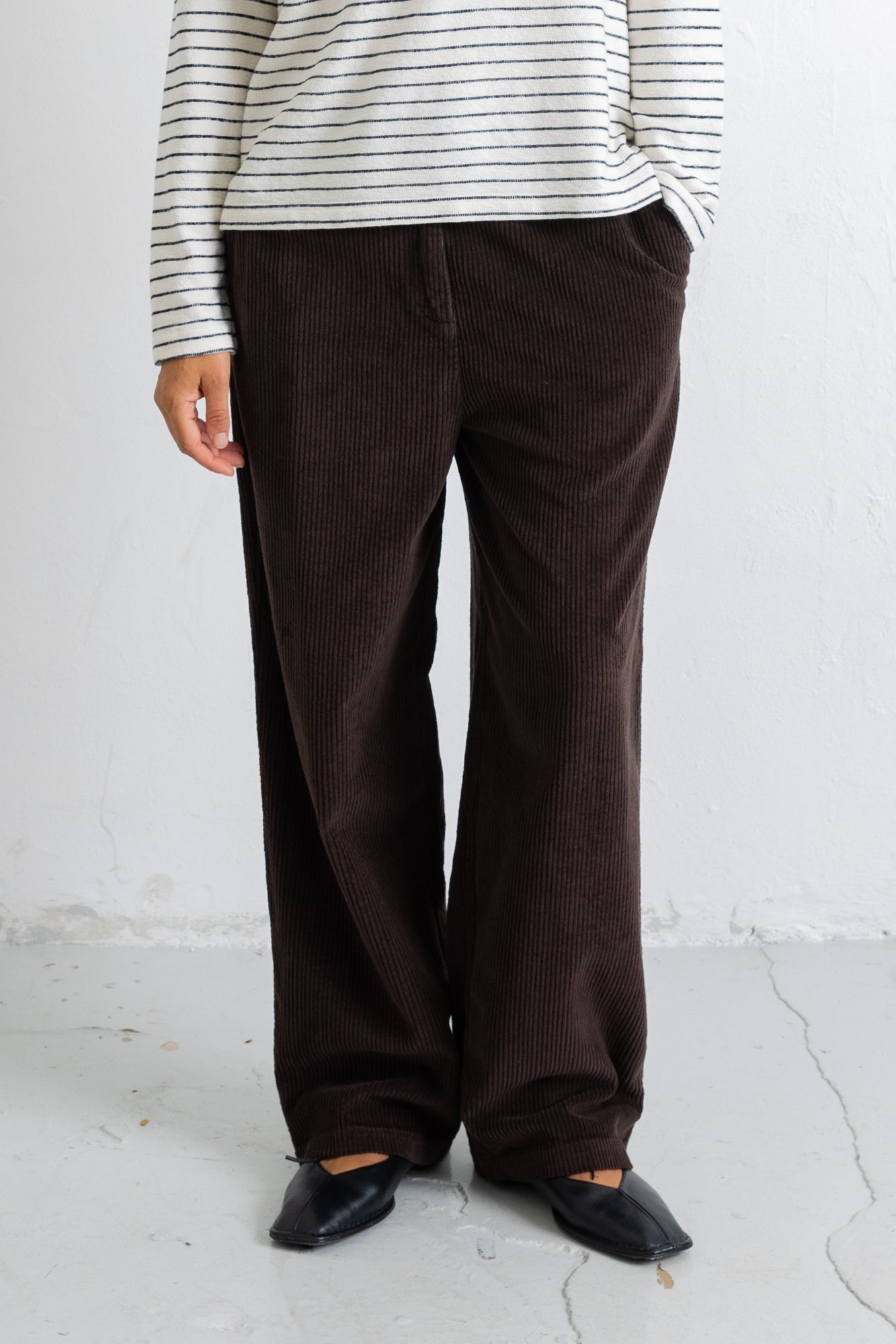 A person wearing dark brown corduroy wide-leg trousers, a cream and navy striped long-sleeve top, and black pointed flats, standing against a white textured wall.