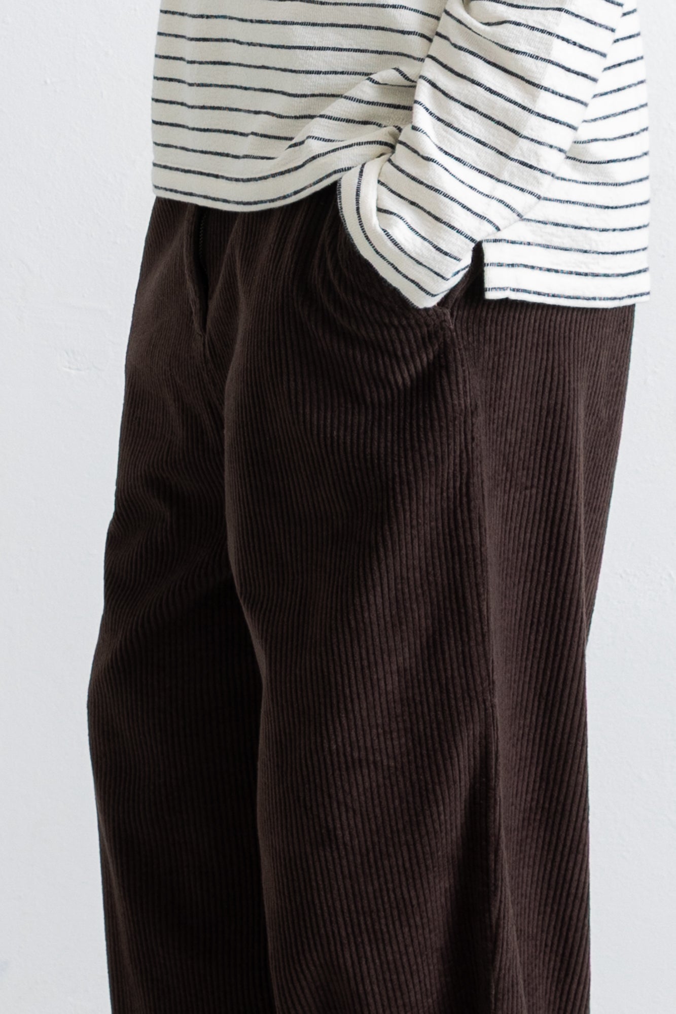 Close-up of a person wearing dark brown corduroy trousers and a white long-sleeve shirt with thin black horizontal stripes, cropped from the chest down against a plain light background.