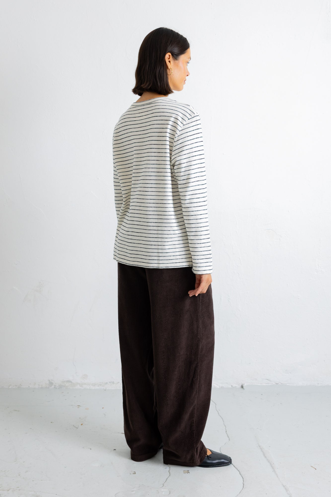 A model shown from the back wearing a cream-colored long-sleeve shirt with thin dark horizontal stripes and dark brown wide-leg trousers, standing against a plain white wall.