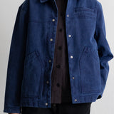 A model wearing a dark indigo blue canvas utility jacket over a black shirt and black pants, standing against a light gray studio background.