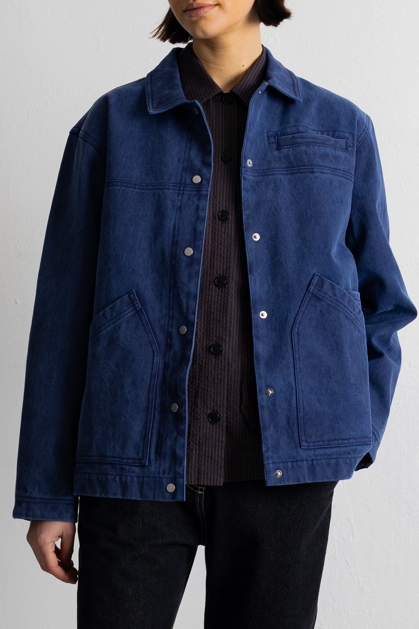 A model wearing a dark indigo blue canvas utility jacket over a black shirt and black pants, standing against a light gray studio background.