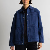 A model wearing a dark indigo blue canvas utility jacket over a black shirt and black pants, standing against a light gray studio background.