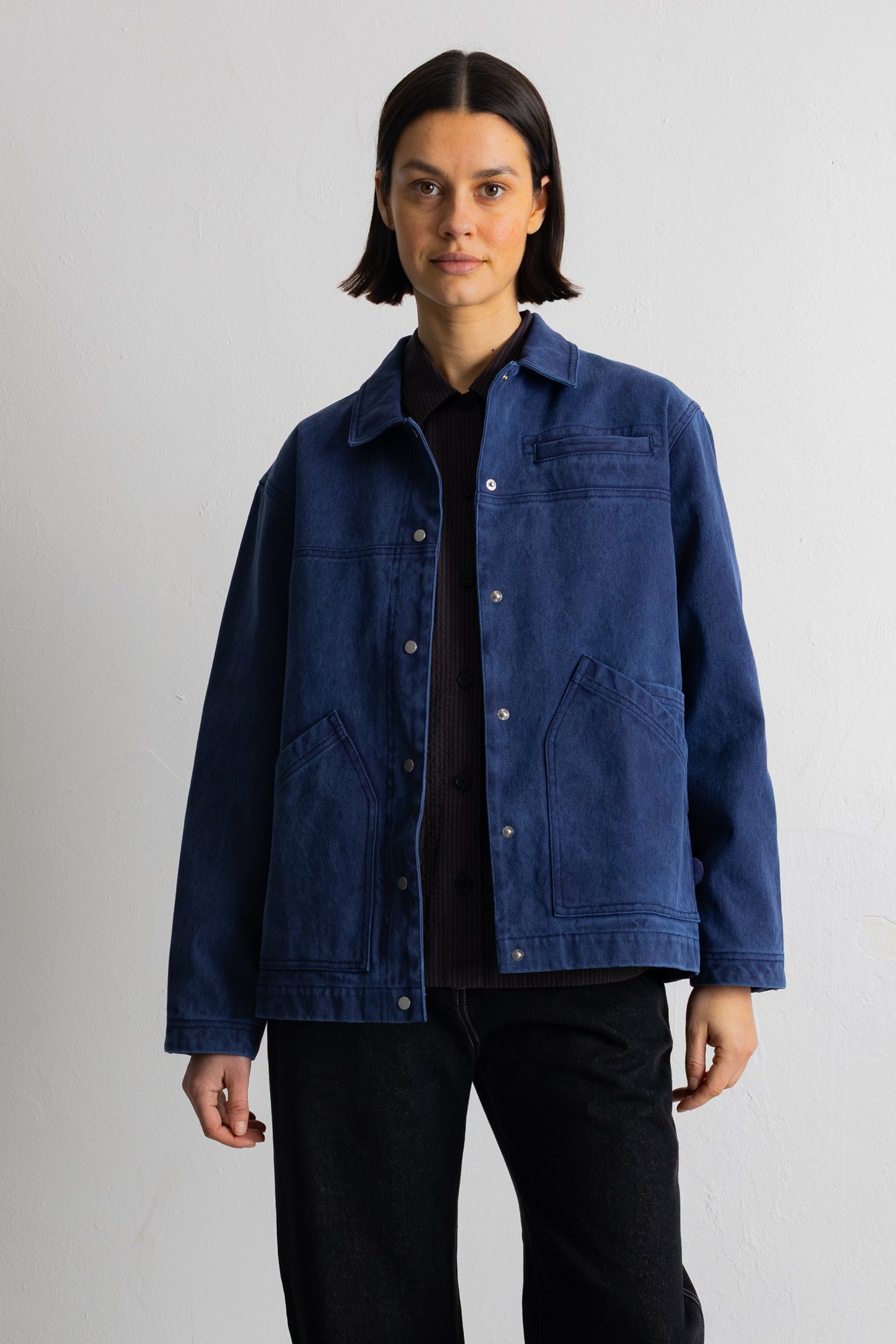 A model wearing a dark indigo blue canvas utility jacket over a black shirt and black pants, standing against a light gray studio background.