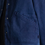 Close-up of a dark blue denim jacket with a large diagonal welt pocket and silver metal snap buttons, worn over a black shirt.