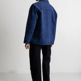 A person shown from the back wearing a dark blue denim jacket, black pants, and black boots, standing against a plain white studio background.