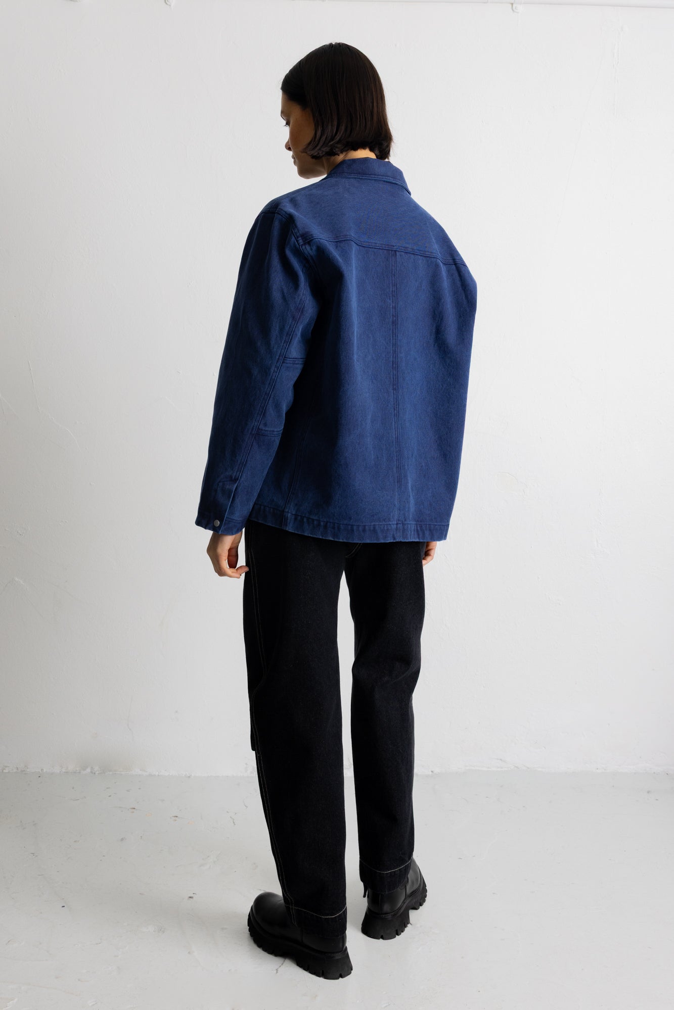 A person shown from the back wearing a dark blue denim jacket, black pants, and black boots, standing against a plain white studio background.