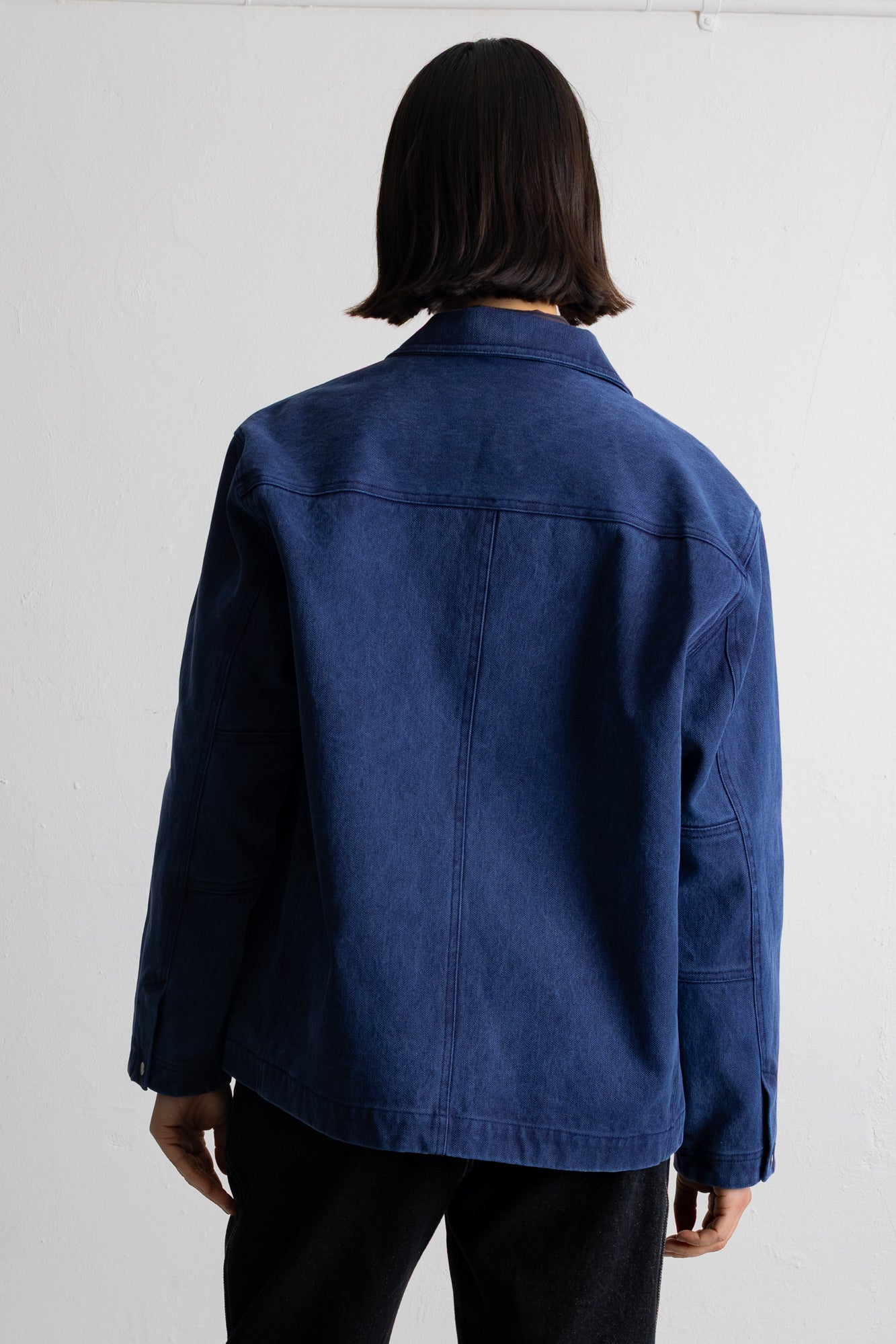 A person shown from the back wearing a dark blue denim jacket and black pants against a plain white wall.