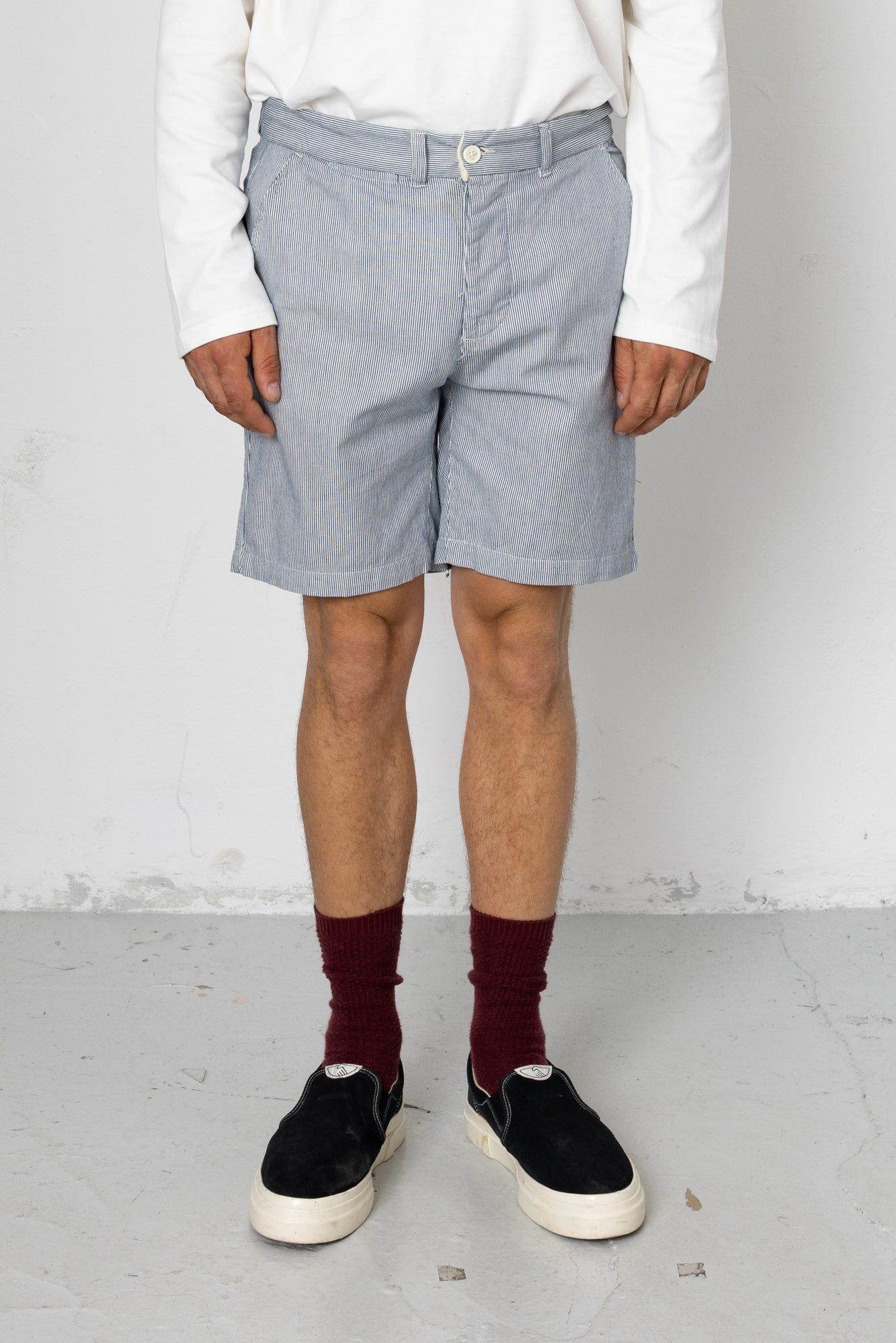 Person wearing light gray checkered shorts, white shirt, maroon socks, and black sneakers on a plain background