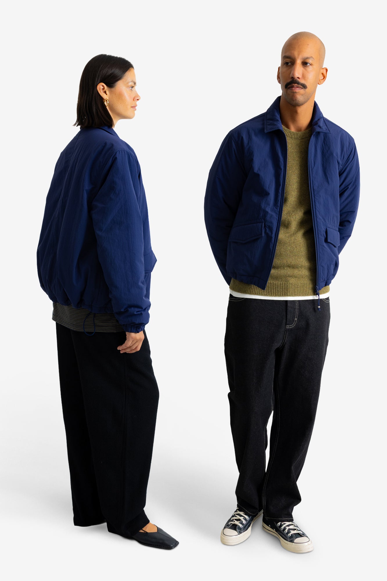 A man and a woman modeling a navy blue lightweight jacket against a clean white studio background. The woman is shown from the back, and the man is shown from the front, both wearing dark pants and sneakers.