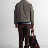 A man viewed from behind wearing an olive green jacket, dark brown corduroy pants, and black shoes, holding a brown and navy patterned tote bag against a white studio wall.