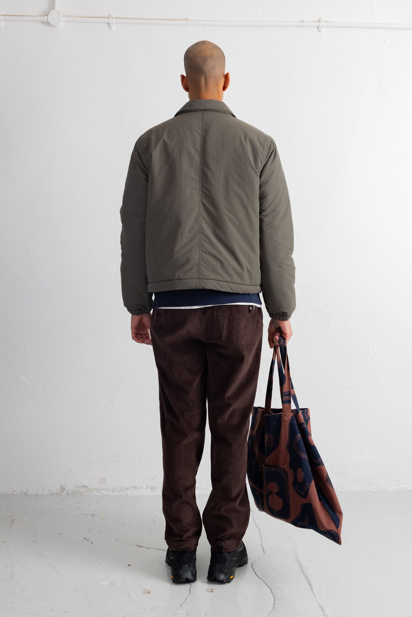 A man viewed from behind wearing an olive green jacket, dark brown corduroy pants, and black shoes, holding a brown and navy patterned tote bag against a white studio wall.
