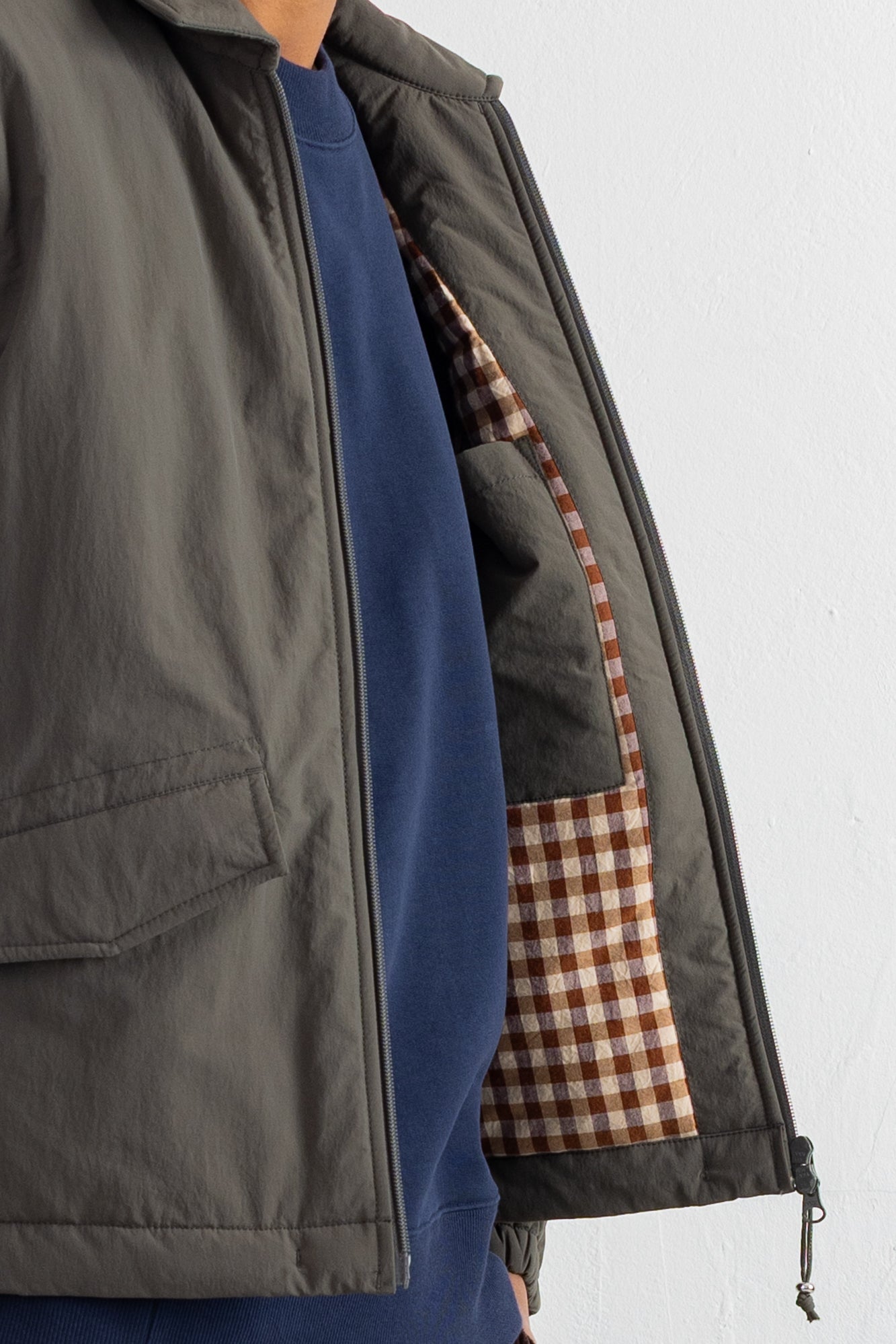 Close-up of a dark gray zip-up jacket worn over a navy blue sweatshirt, showing the interior pocket lined with a brown and beige gingham pattern.