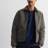 A man wearing a dark olive green zip-up puffer jacket over a navy blue crewneck sweatshirt and brown corduroy trousers, standing against a plain white wall.
