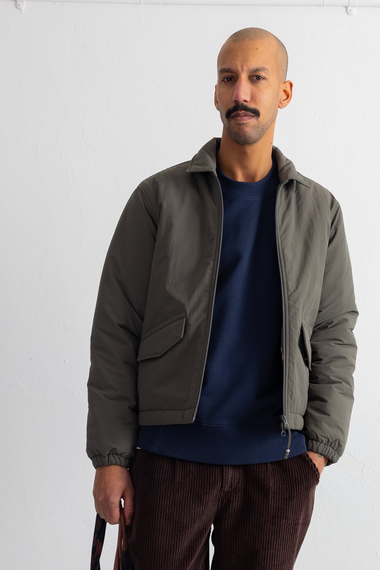 A man wearing a dark olive green zip-up puffer jacket over a navy blue crewneck sweatshirt and brown corduroy trousers, standing against a plain white wall.