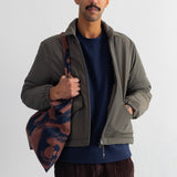 A man wearing an olive green zip-up jacket over a navy blue shirt and brown corduroy pants, holding a brown and navy patterned shoulder bag against a plain white wall.