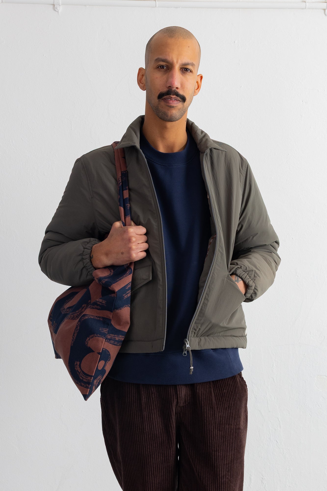 A man wearing an olive green zip-up jacket over a navy blue shirt and brown corduroy pants, holding a brown and navy patterned shoulder bag against a plain white wall.
