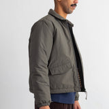 A man shown in profile wearing an olive green puffer jacket over a blue shirt and brown corduroy pants, standing against a plain white wall.