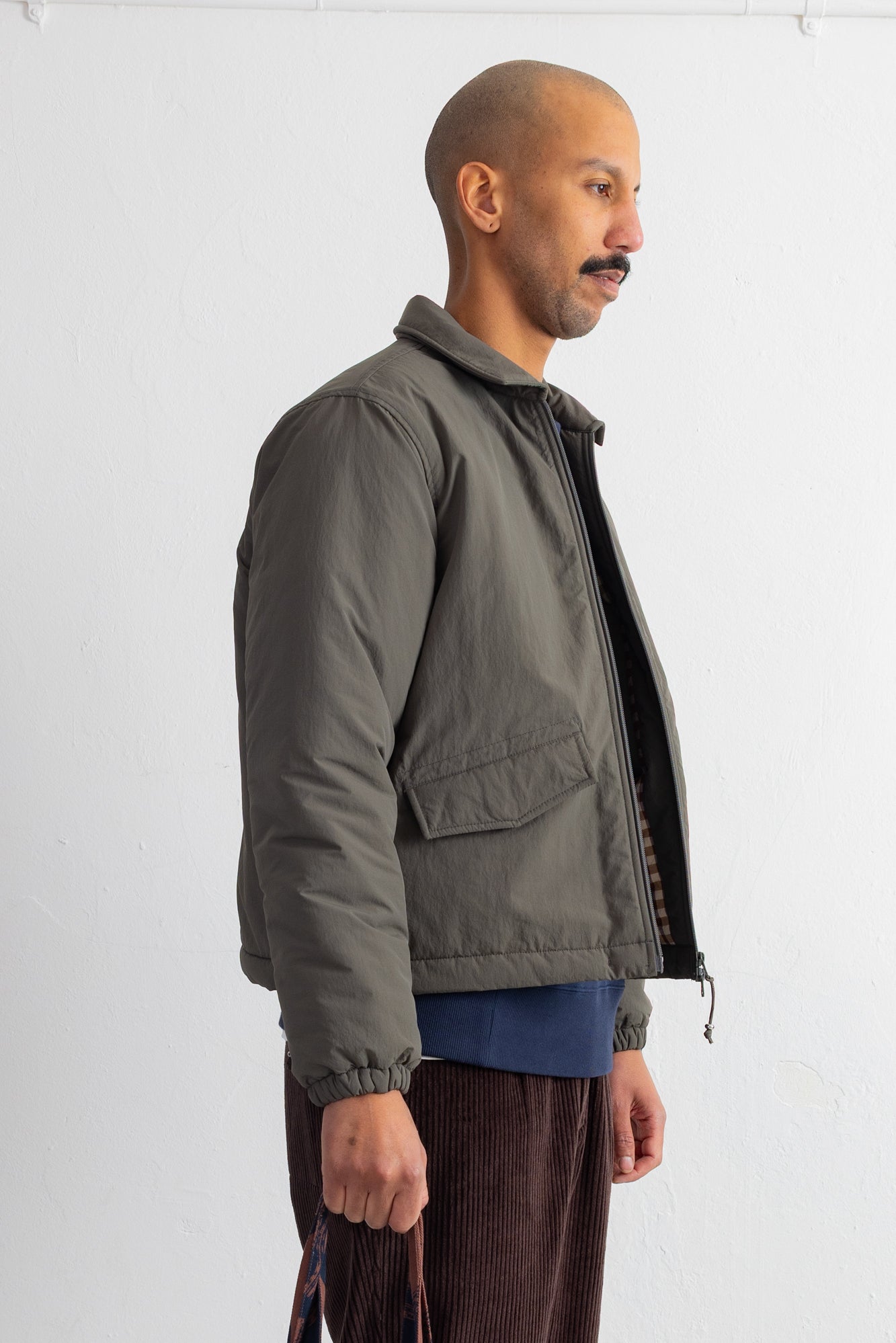 A man shown in profile wearing an olive green puffer jacket over a blue shirt and brown corduroy pants, standing against a plain white wall.
