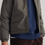 A close-up of a person wearing an olive green lightweight zip-front jacket over a navy blue crewneck sweatshirt, paired with dark brown corduroy trousers.