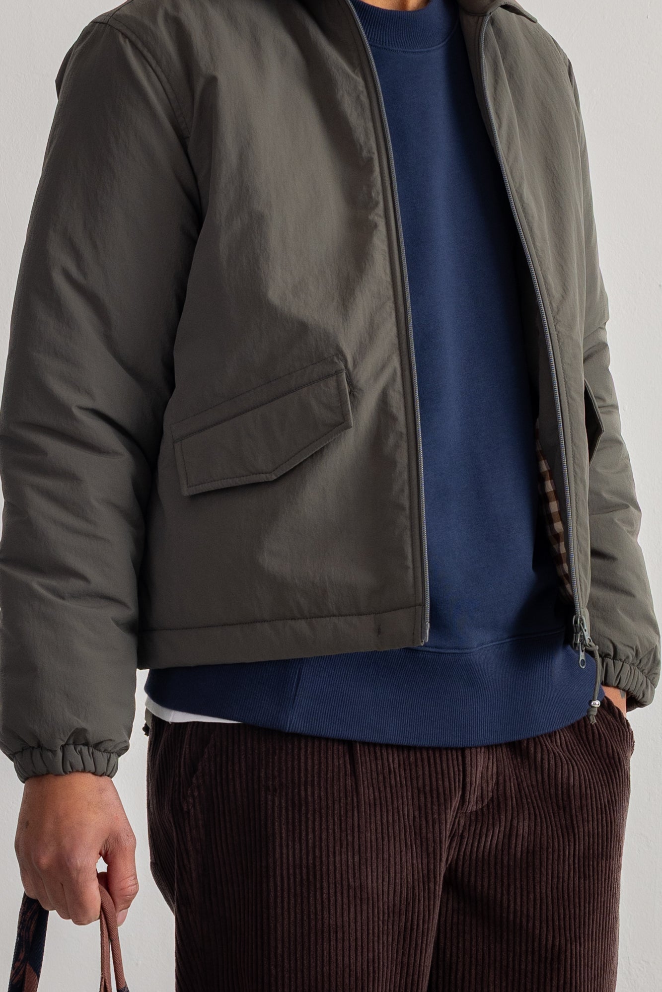 A close-up of a person wearing an olive green lightweight zip-front jacket over a navy blue crewneck sweatshirt, paired with dark brown corduroy trousers.
