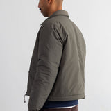 A man shown from the side profile wearing a dark olive green puffer jacket, a navy blue shirt, and brown corduroy trousers, standing against a white wall.