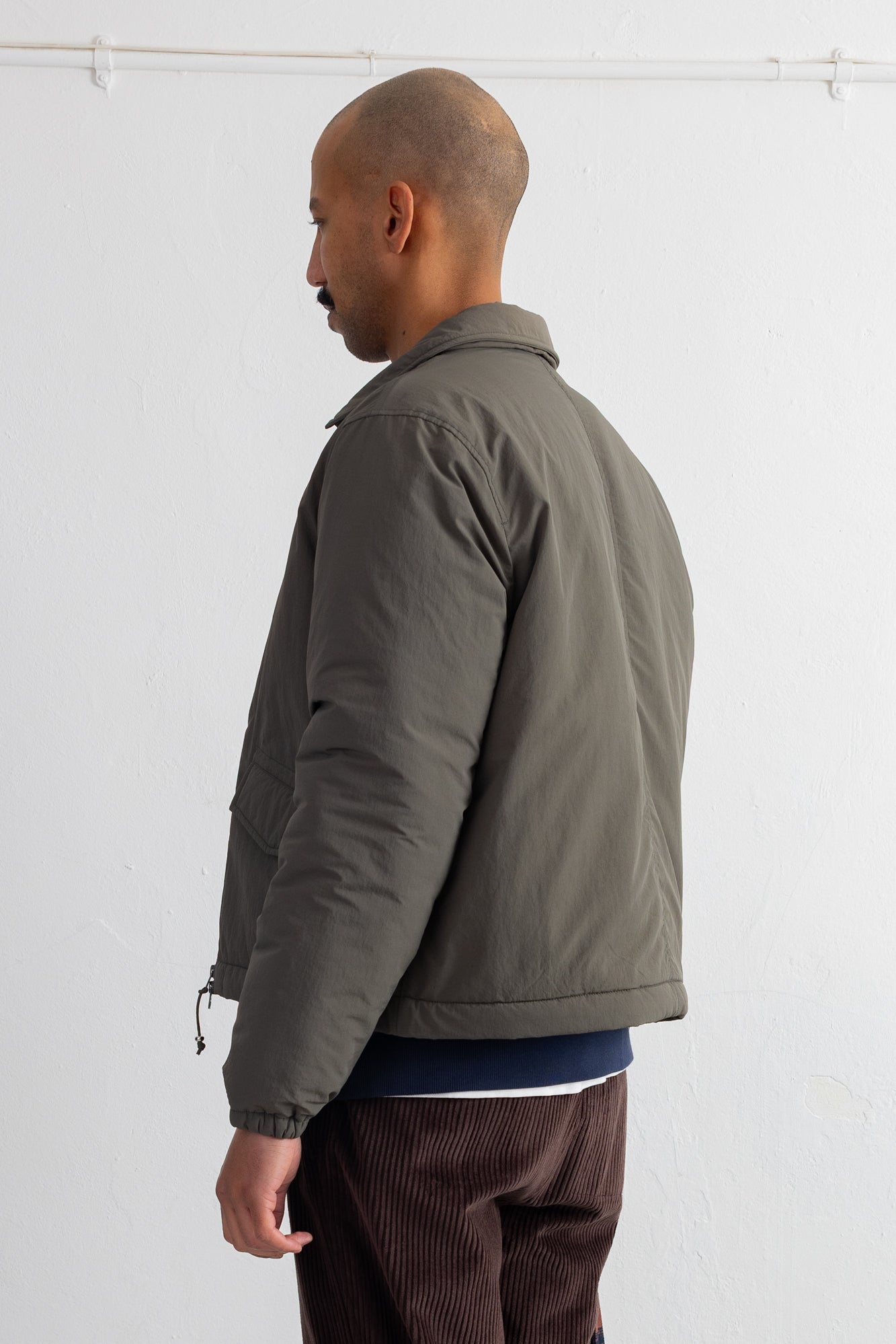 A man shown from the side profile wearing a dark olive green puffer jacket, a navy blue shirt, and brown corduroy trousers, standing against a white wall.