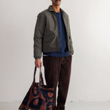 A man stands against a white wall wearing a dark olive jacket, navy shirt, brown corduroy pants, and black shoes, while holding a brown tote bag with a dark blue abstract pattern.
