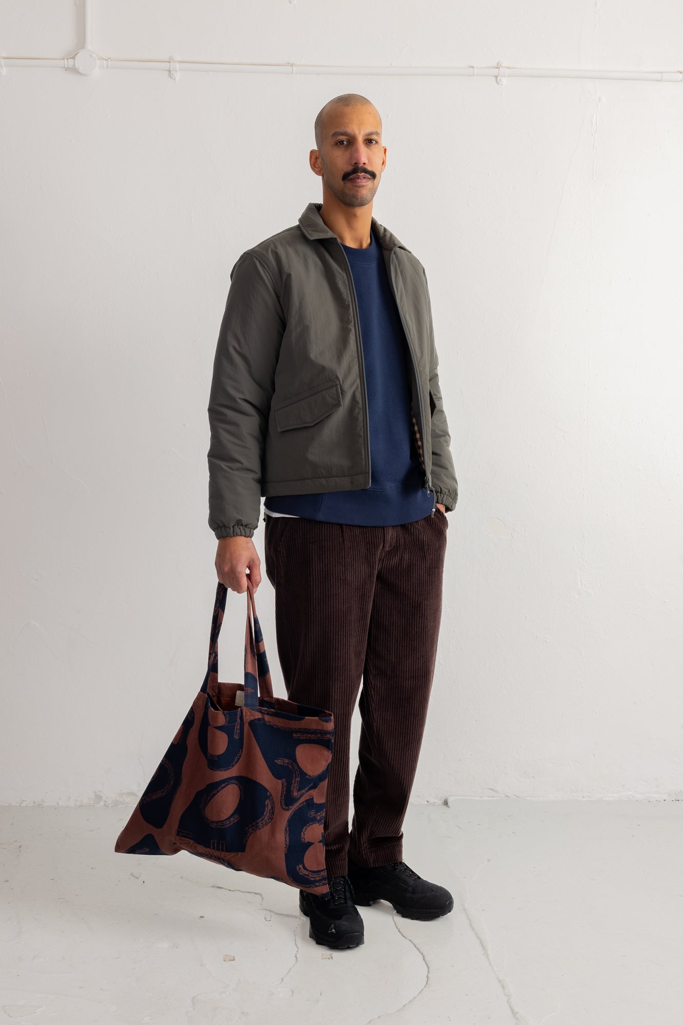 A man stands against a white wall wearing a dark olive jacket, navy shirt, brown corduroy pants, and black shoes, while holding a brown tote bag with a dark blue abstract pattern.