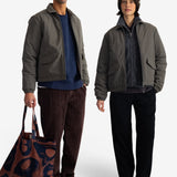 Two models wearing matching dark olive green puffer jackets, standing against a plain white studio background. The model on the left wears brown corduroy trousers and holds a brown and navy patterned tote bag, while the model on the right wears black trousers and a brown knit beanie.