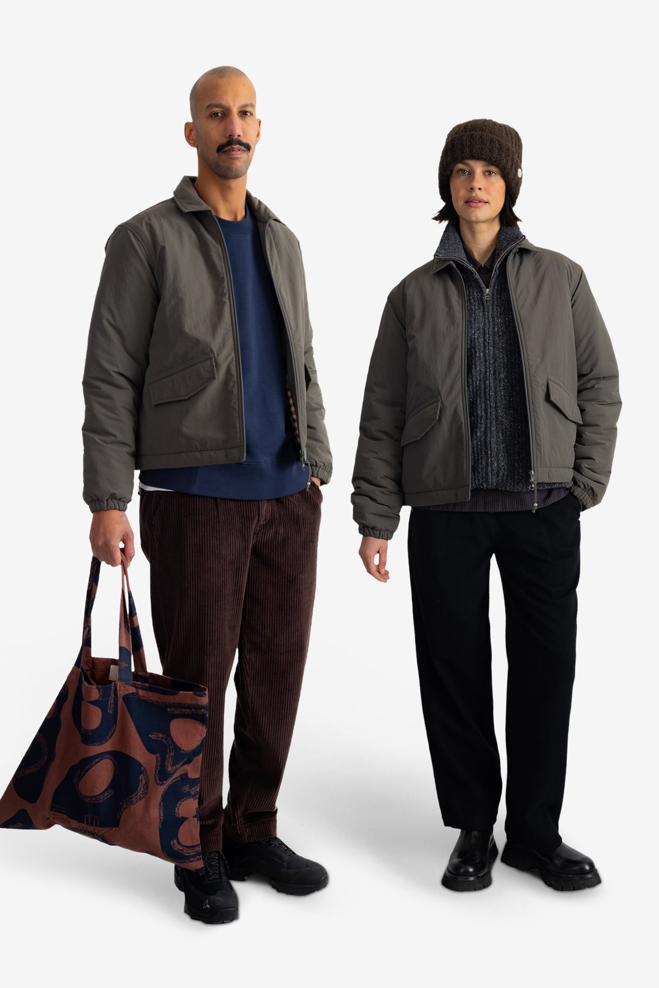 Two models wearing matching dark olive green puffer jackets, standing against a plain white studio background. The model on the left wears brown corduroy trousers and holds a brown and navy patterned tote bag, while the model on the right wears black trousers and a brown knit beanie.