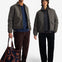 Two models wearing matching dark olive green puffer jackets, standing against a plain white studio background. The model on the left wears brown corduroy trousers and holds a brown and navy patterned tote bag, while the model on the right wears black trousers and a brown knit beanie.