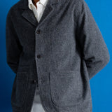 A man wearing a dark gray textured wool-blend jacket over a white collared shirt, standing against a solid blue background.