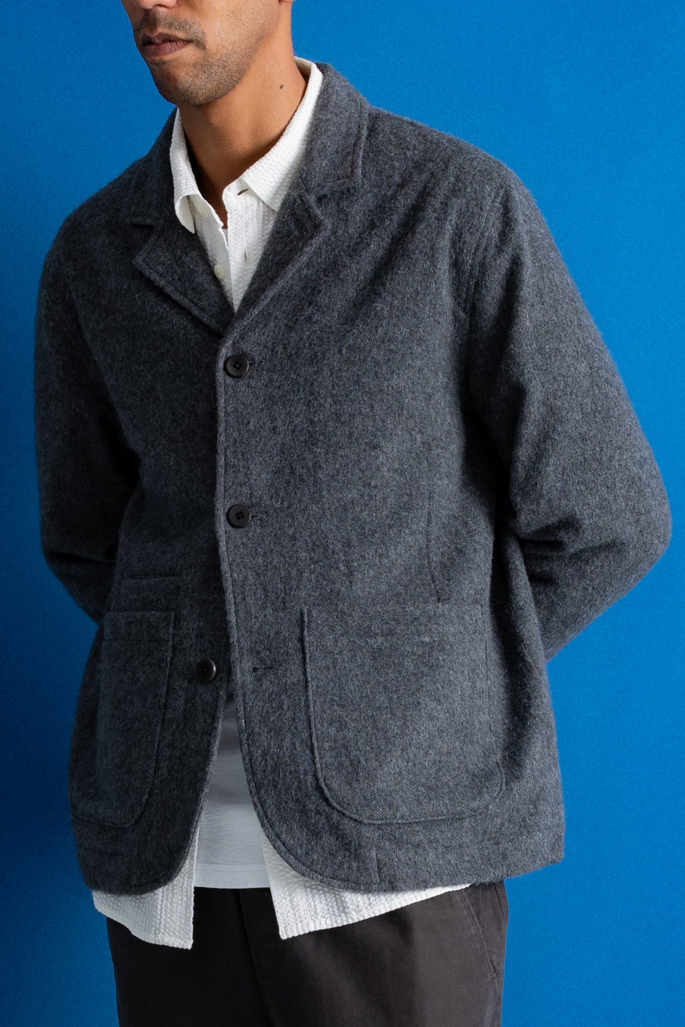 A man wearing a dark gray textured wool-blend jacket over a white collared shirt, standing against a solid blue background.