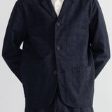 A man wearing a dark grey textured linen-blend blazer jacket over a white collared shirt, paired with matching navy trousers, shot against a light gray background.