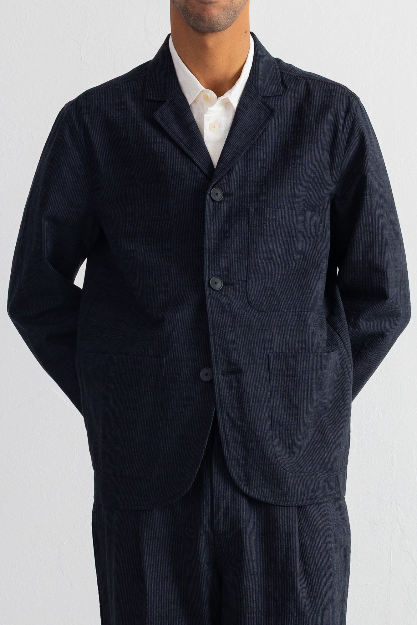A man wearing a dark grey textured linen-blend blazer jacket over a white collared shirt, paired with matching navy trousers, shot against a light gray background.