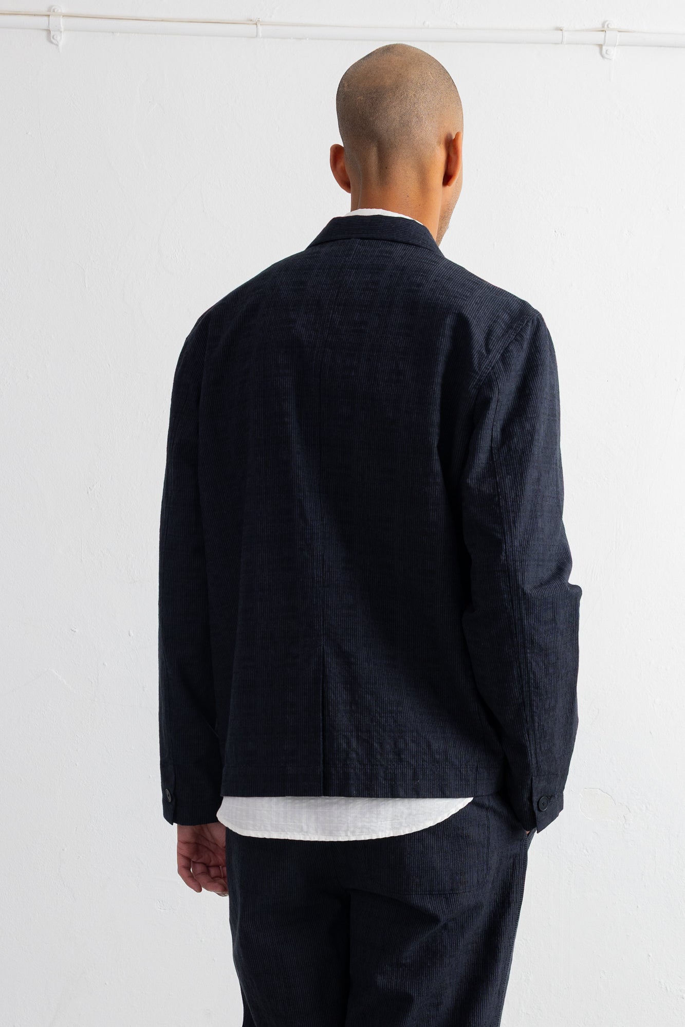 A man viewed from behind wearing a dark grey textured jacket over a white shirt and matching dark trousers, standing against a plain white wall.