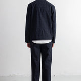 A man viewed from behind wearing a dark grey jacket and matching trousers, layered over a white shirt, standing against a white studio wall.