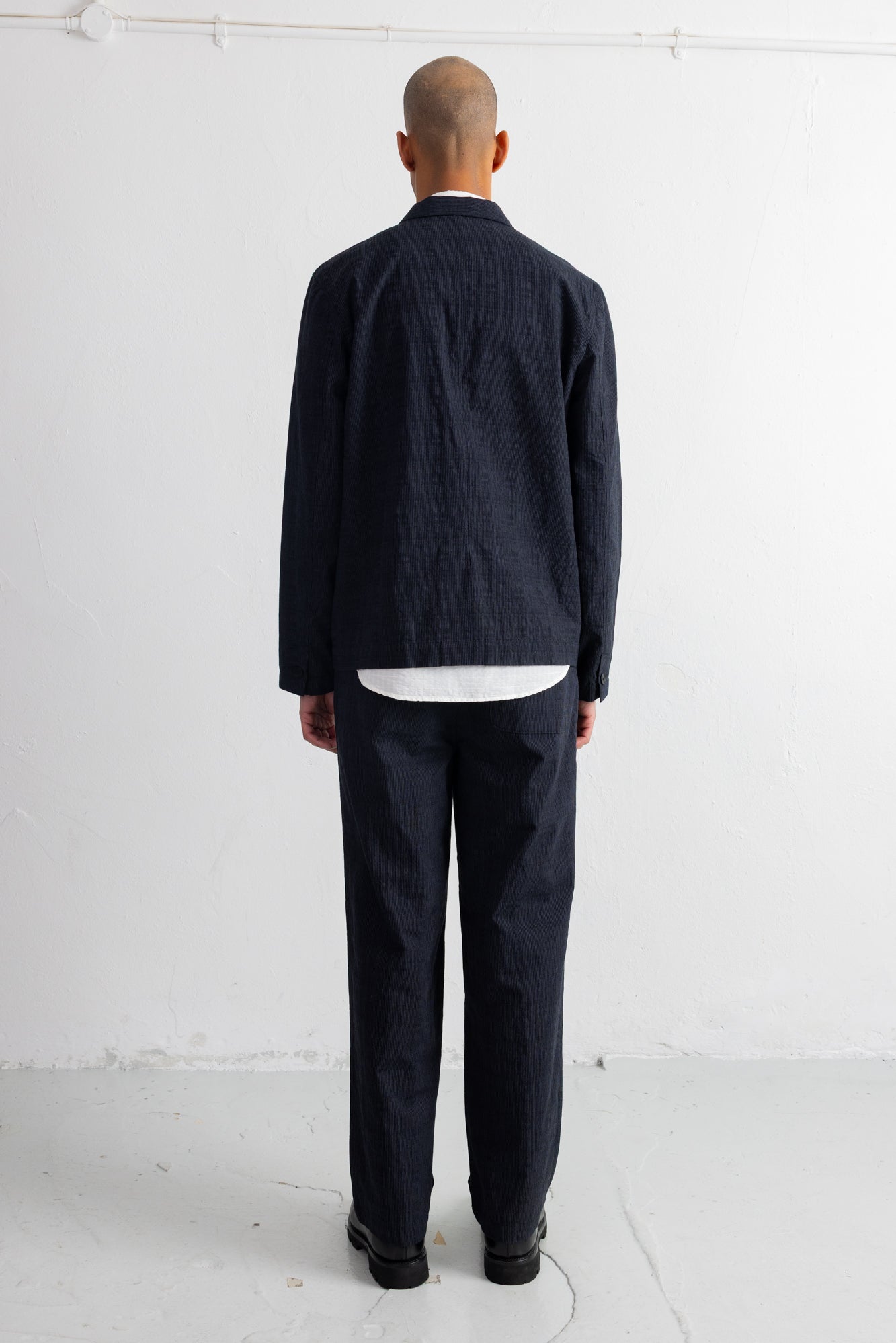 A man viewed from behind wearing a dark grey jacket and matching trousers, layered over a white shirt, standing against a white studio wall.
