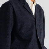 A close-up of a man wearing a dark grey corduroy blazer over a cream-colored collared shirt, against a plain light background.