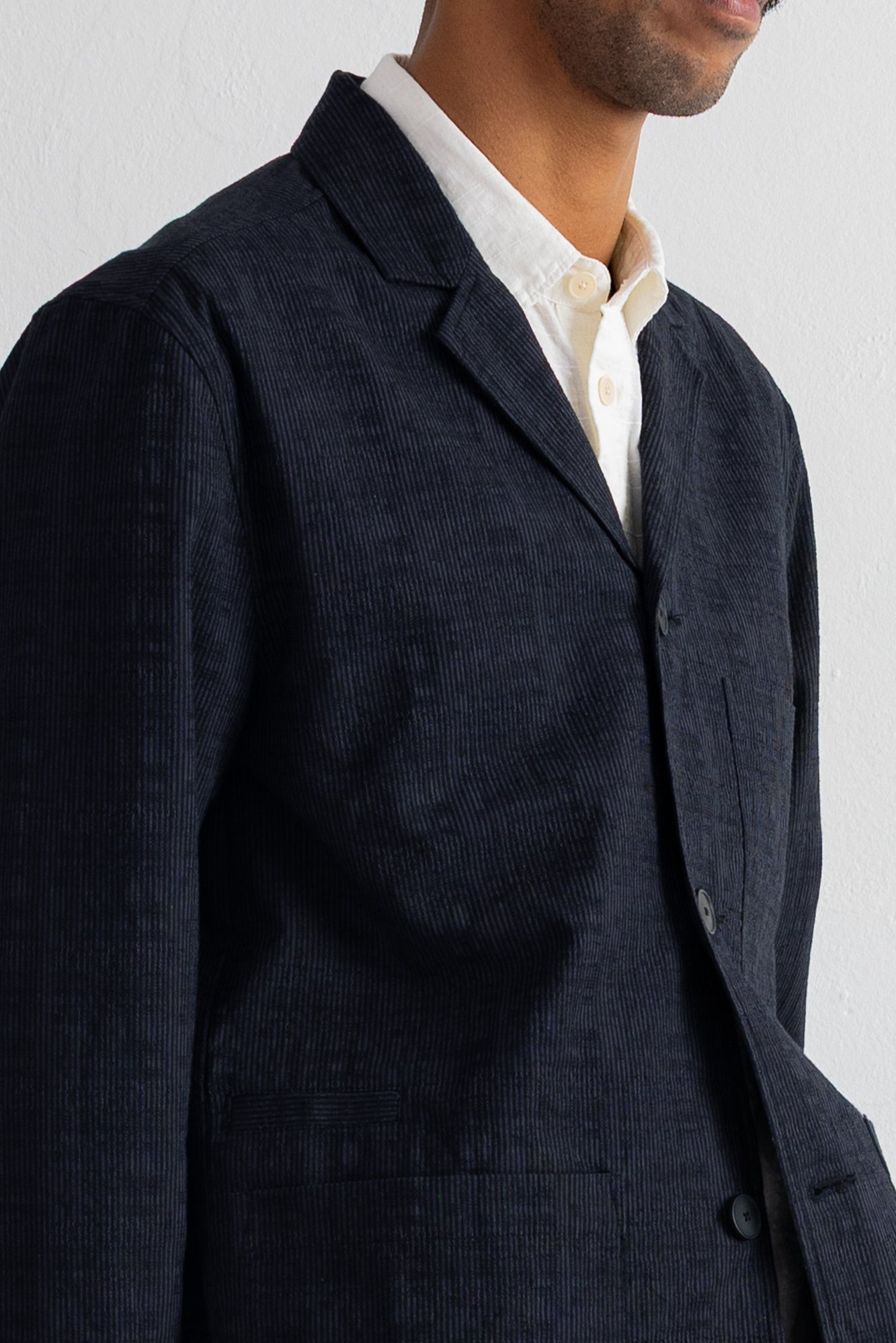 A close-up of a man wearing a dark grey corduroy blazer over a cream-colored collared shirt, against a plain light background.