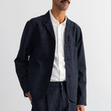 A man wearing a dark grey blazer and matching trousers over a white button-down shirt, standing against a white wall.