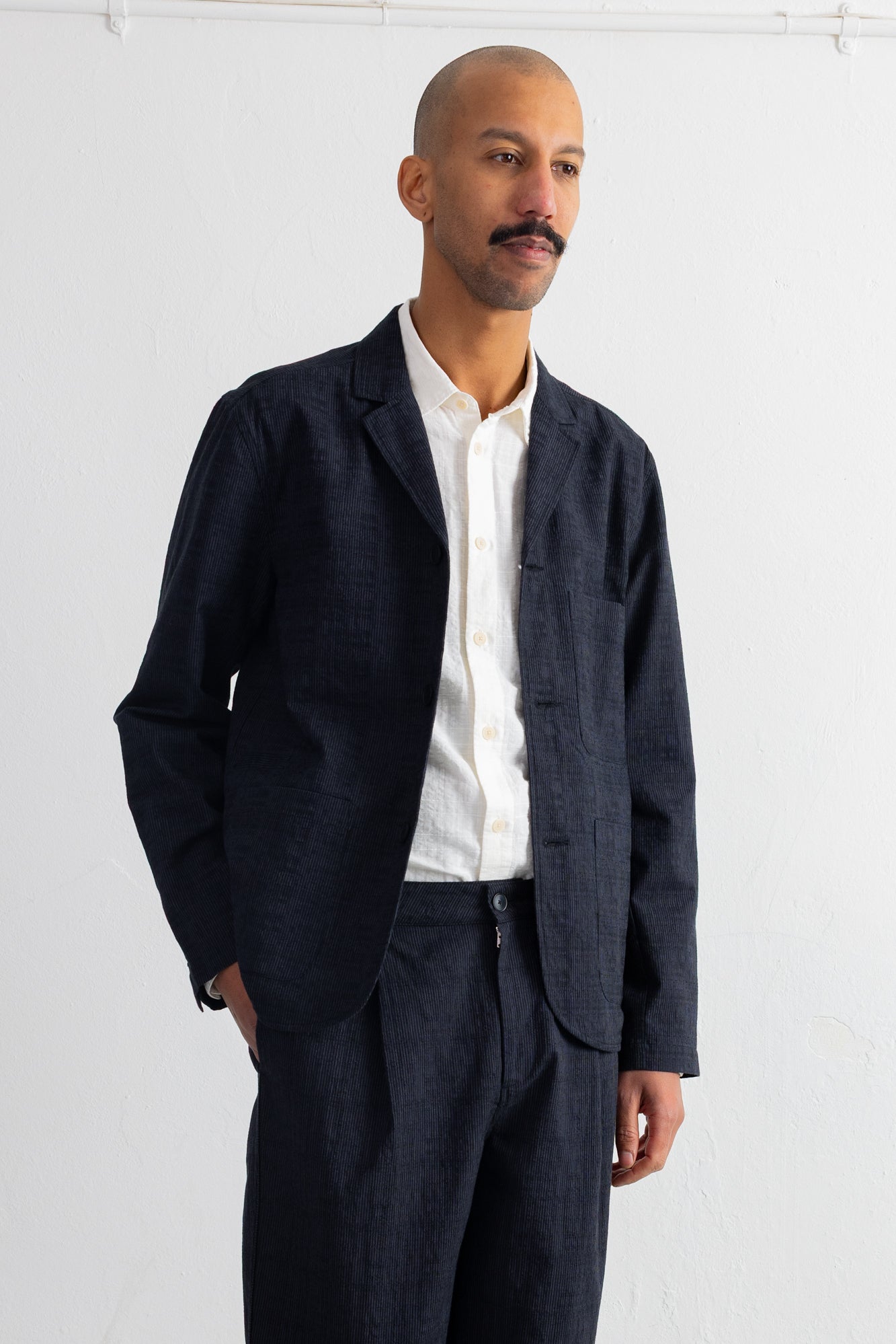 A man wearing a dark grey blazer and matching trousers over a white button-down shirt, standing against a white wall.