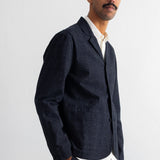 A man wearing a dark charcoal linen-blend blazer over a white collared shirt, standing against a plain white wall.