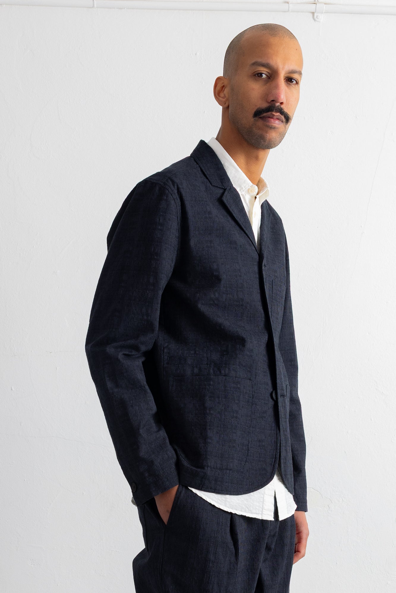 A man wearing a dark charcoal linen-blend blazer over a white collared shirt, standing against a plain white wall.