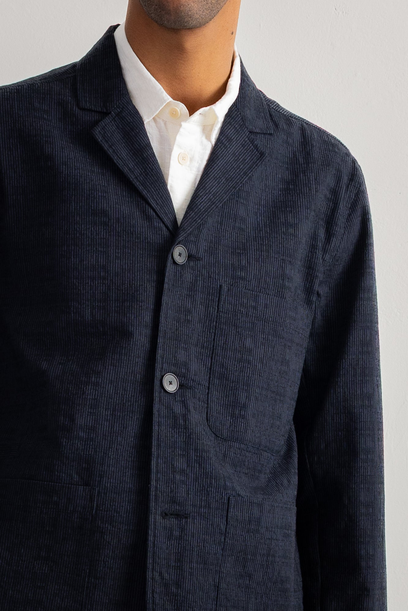 A close-up of a man wearing a dark greytextured jacket over a white collared shirt, cropped from the chin down against a plain light background.