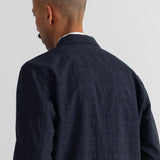 A man viewed from behind wearing a dark grey jacket over a white collared shirt, set against a plain white wall.