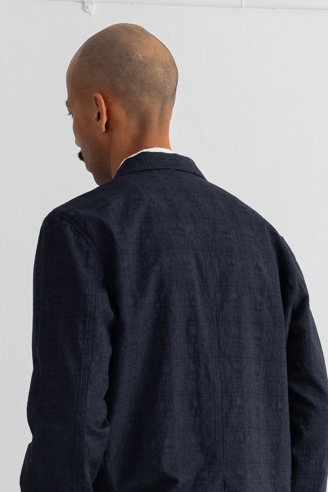 A man viewed from behind wearing a dark grey jacket over a white collared shirt, set against a plain white wall.