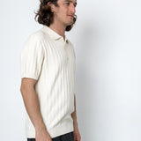 Man wearing a white knit polo shirt and dark pants against a white background