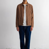 A man wearing a brown suede jacket over a white striped t-shirt, dark blue jeans, black shoes, and a navy blue beanie, standing against a white wall.